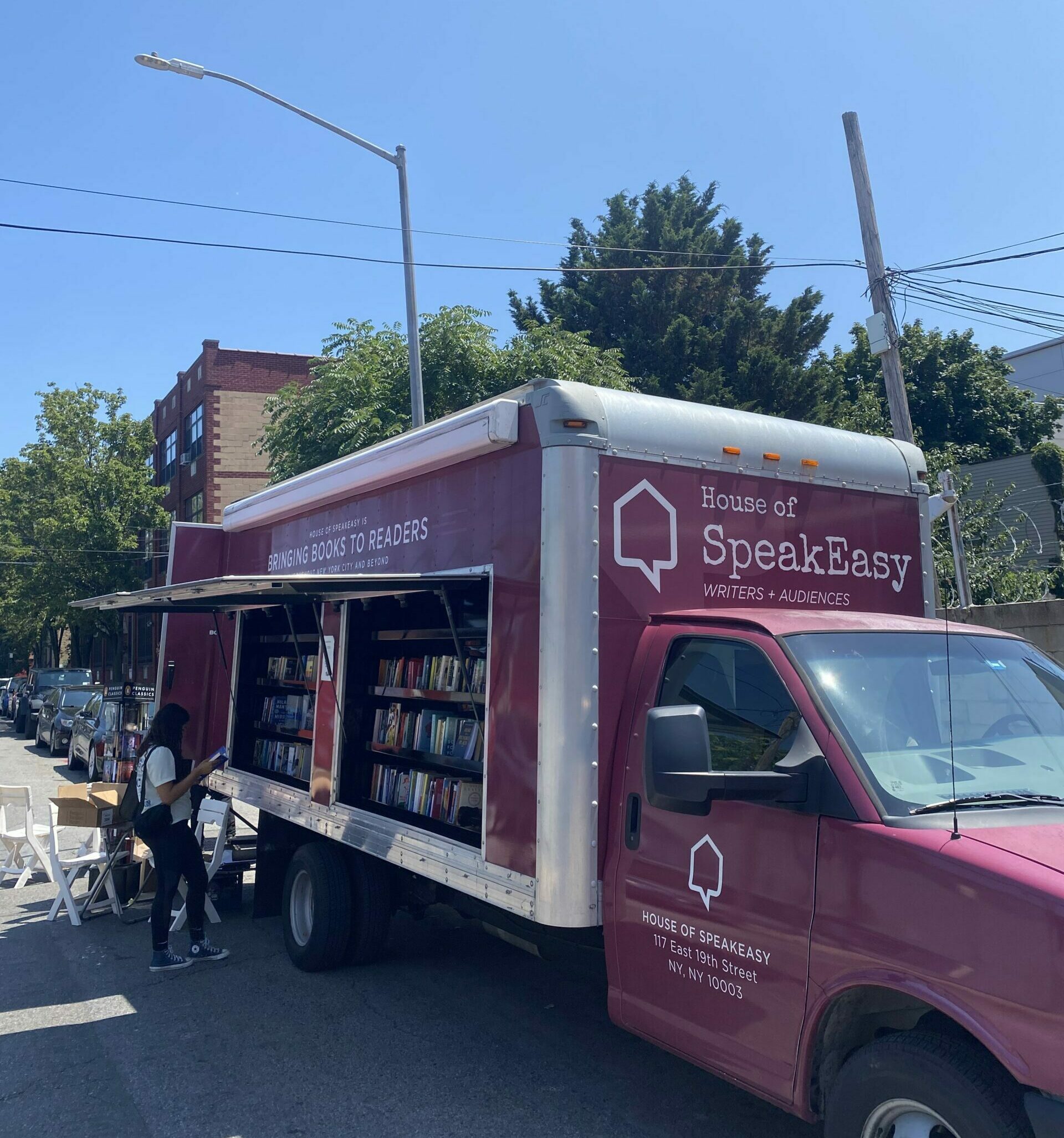 SpeakEasy's Bookmobile on a sunny, open street. Someone stands in the shade under the truck's awning, looking at the books on offer.