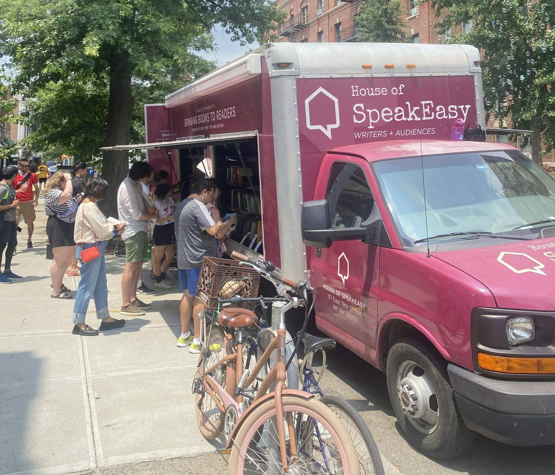 House of SpeakEasy's Bookmobile, parked on a sunny street behind a rack of bikes. A crowd of people is gathered by the Bookmobile's shelves, choosing their books.