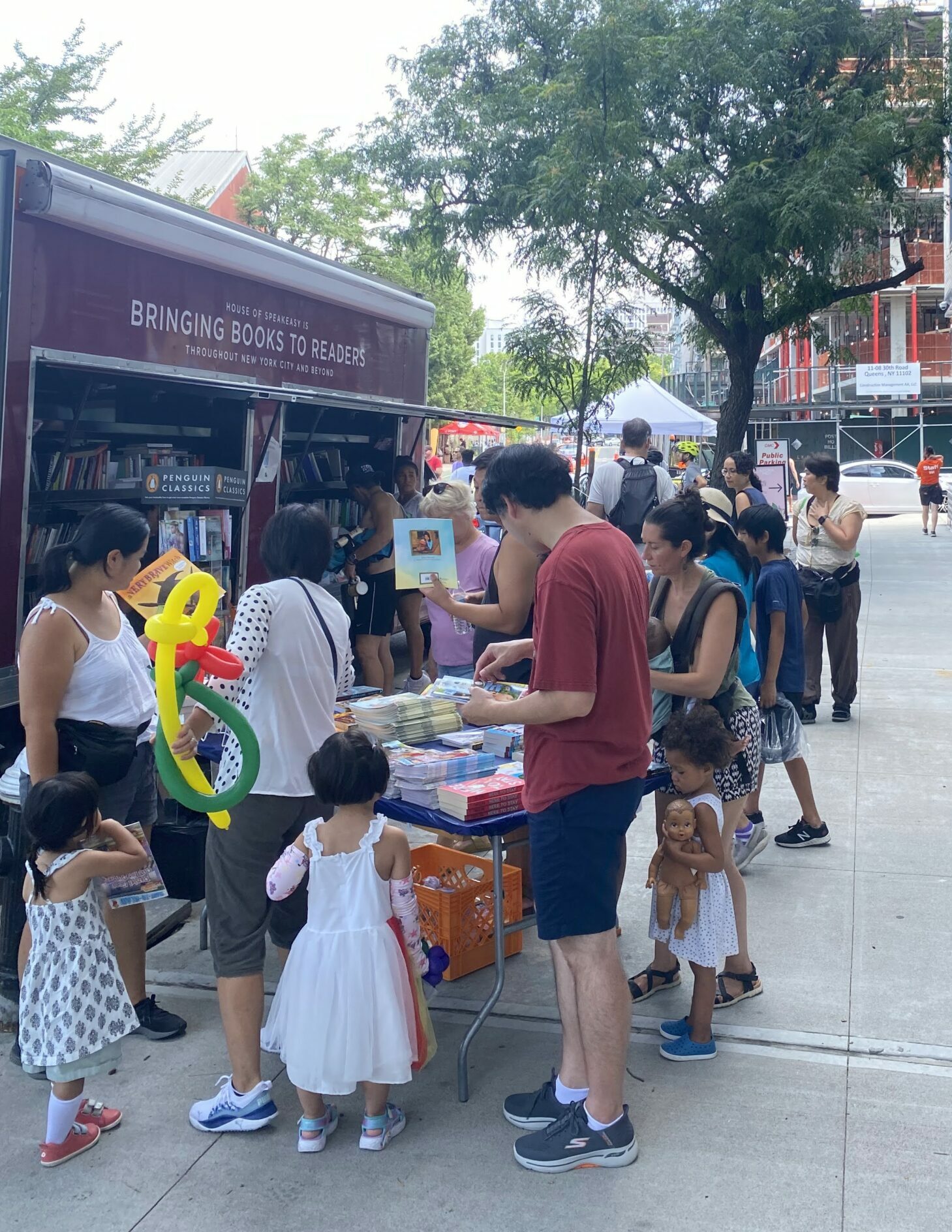 House of SpeakEasy's Bookmobile, parked on a sunny street behind a rack of bikes. A crowd of people is gathered by the Bookmobile's shelves, choosing their books.