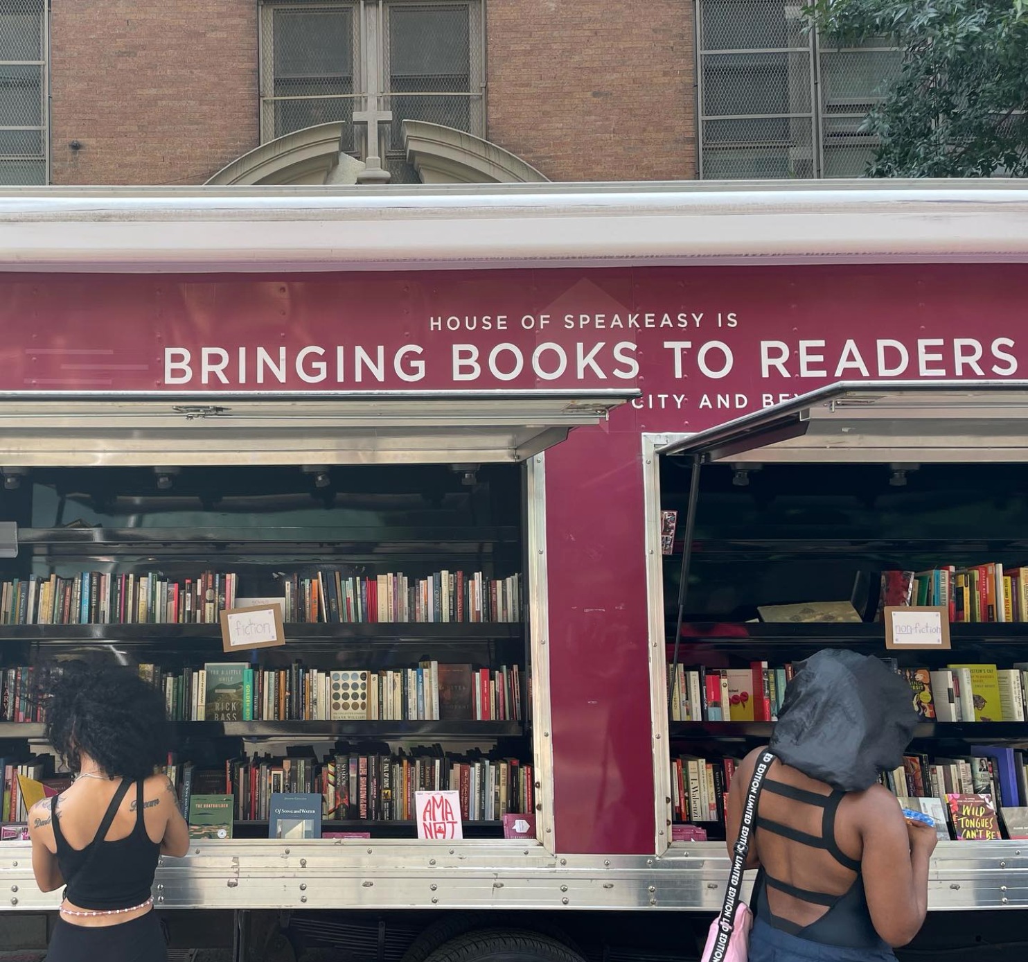 House of SpeakEasy's Bookmobile, parked on a sunny street behind a rack of bikes. A crowd of people is gathered by the Bookmobile's shelves, choosing their books.