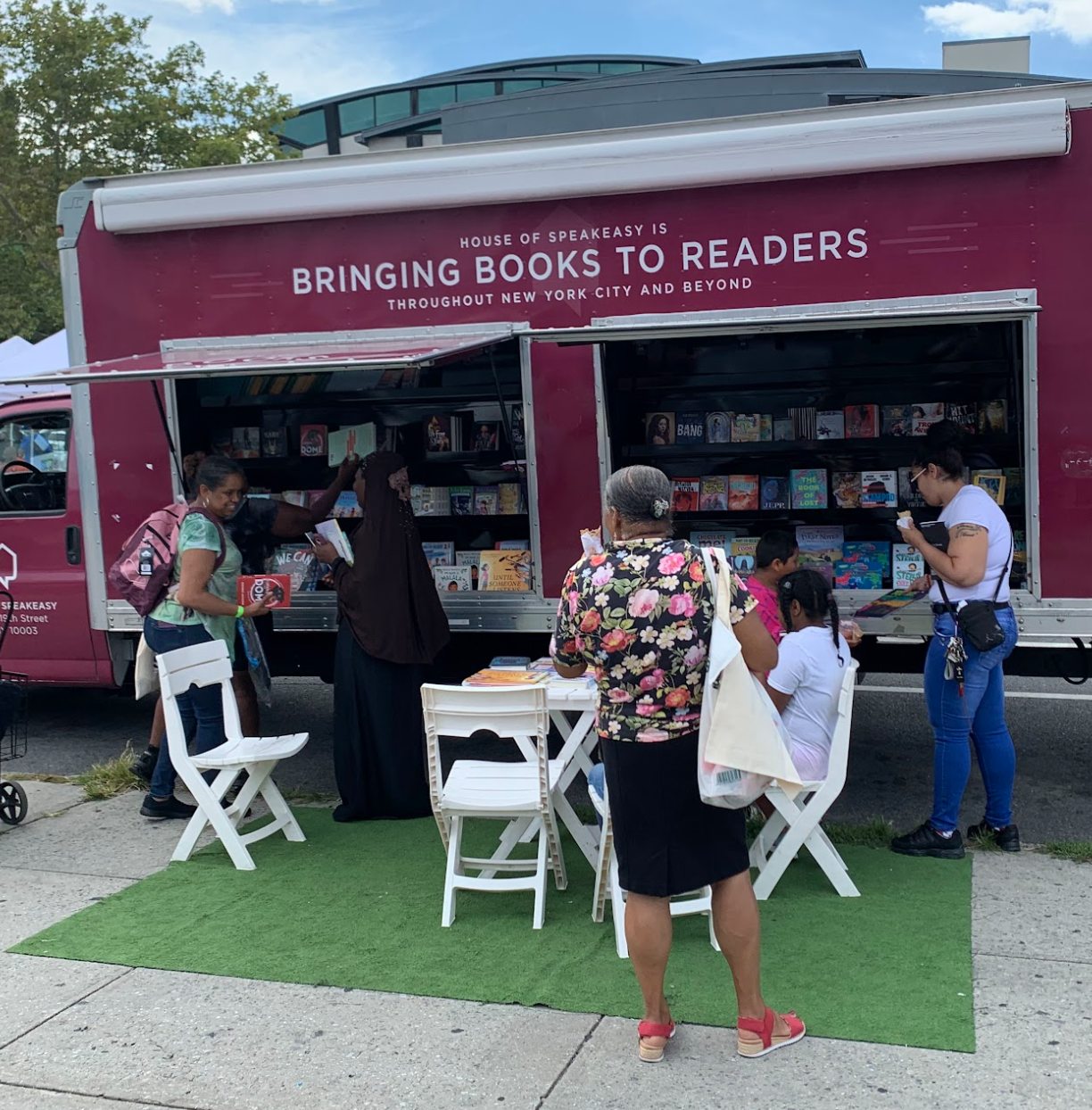 House of SpeakEasy's Bookmobile, parked on a sunny street behind a rack of bikes. A crowd of people is gathered by the Bookmobile's shelves, choosing their books.