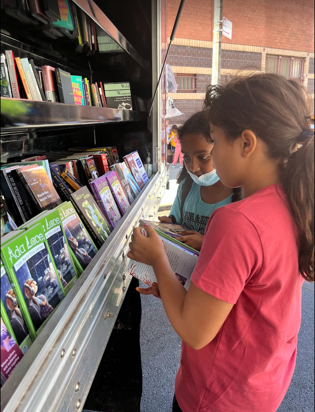 House of SpeakEasy's Bookmobile, parked on a sunny street behind a rack of bikes. A crowd of people is gathered by the Bookmobile's shelves, choosing their books.