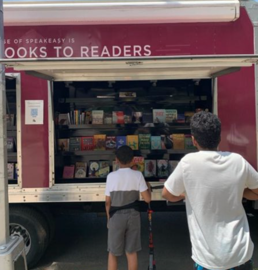 House of SpeakEasy's Bookmobile, parked on a sunny street behind a rack of bikes. A crowd of people is gathered by the Bookmobile's shelves, choosing their books.