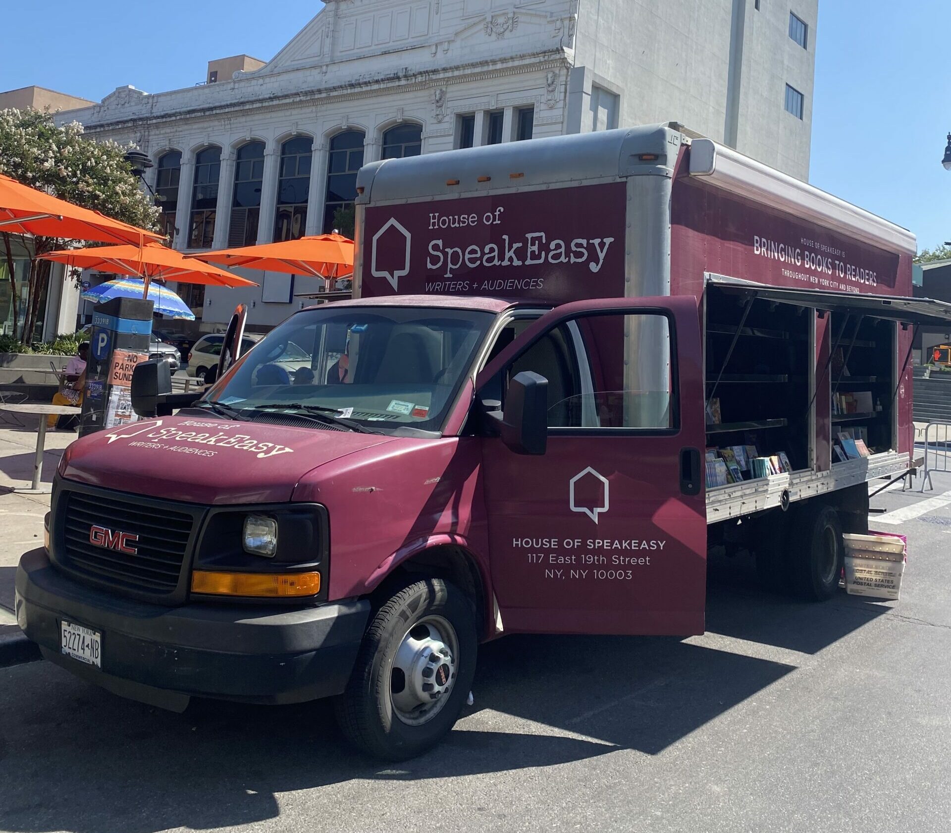 House of SpeakEasy's Bookmobile, parked on a sunny street behind a rack of bikes. A crowd of people is gathered by the Bookmobile's shelves, choosing their books.