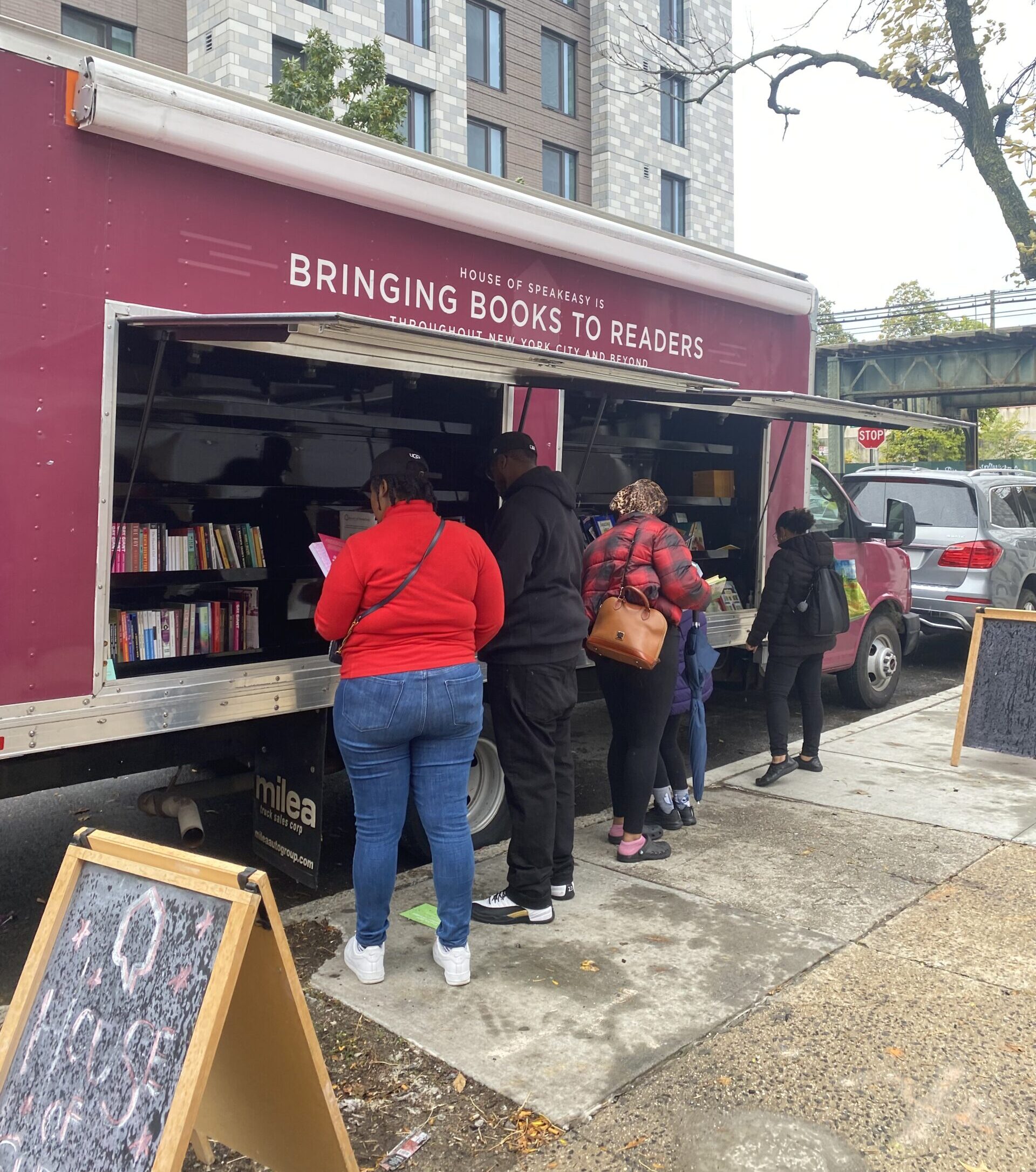 House of SpeakEasy's Bookmobile, parked on a sunny street behind a rack of bikes. A crowd of people is gathered by the Bookmobile's shelves, choosing their books.