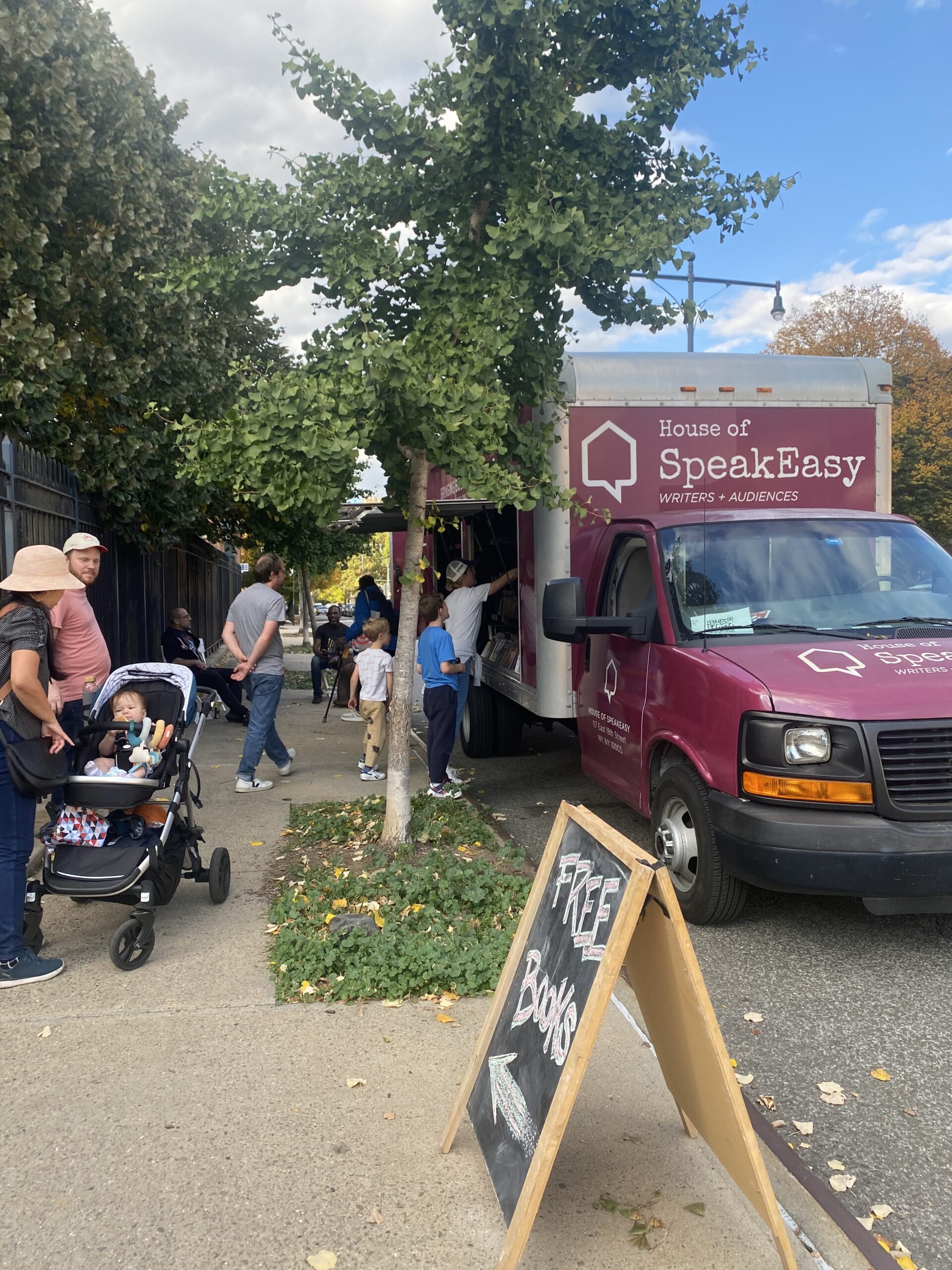 House of SpeakEasy's Bookmobile, parked on a sunny street behind a rack of bikes. A crowd of people is gathered by the Bookmobile's shelves, choosing their books.