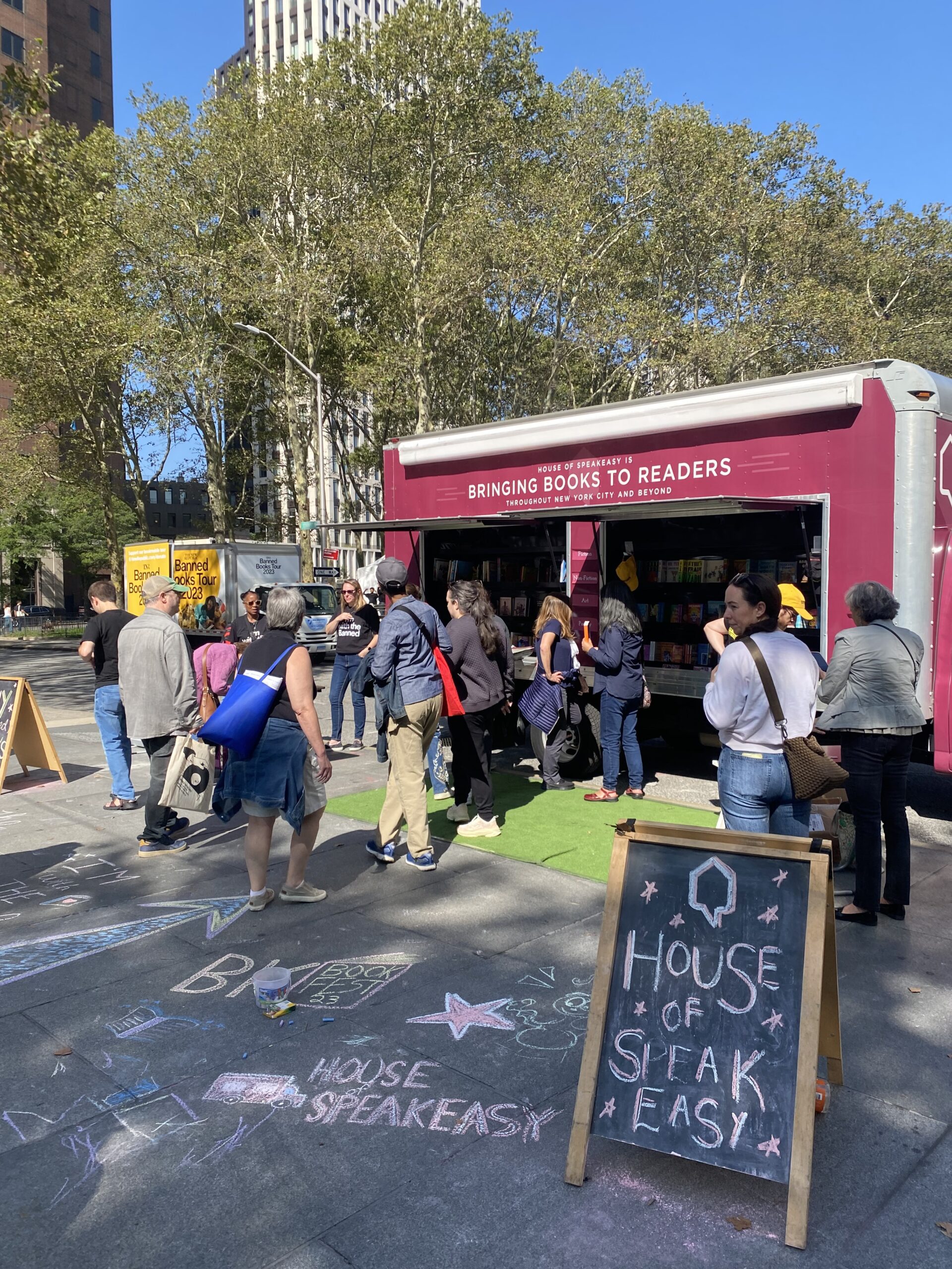 House of SpeakEasy's Bookmobile, parked on a sunny street behind a rack of bikes. A crowd of people is gathered by the Bookmobile's shelves, choosing their books.