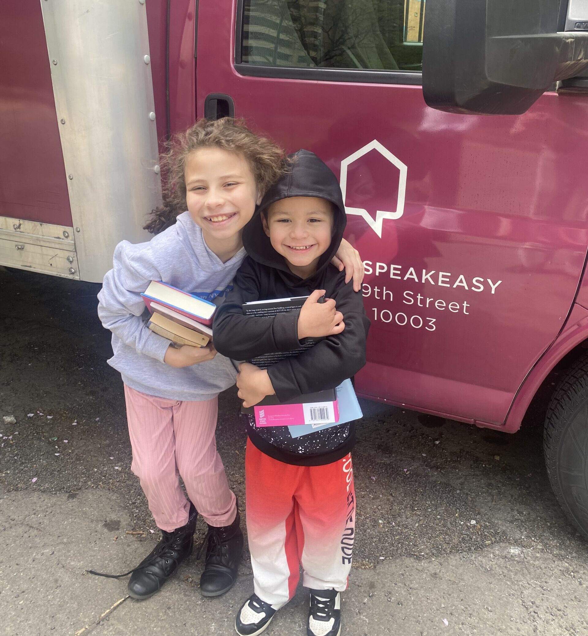 House of SpeakEasy's Bookmobile, parked on a sunny street behind a rack of bikes. A crowd of people is gathered by the Bookmobile's shelves, choosing their books.